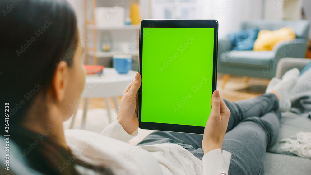 Poster young woman at home resting on a couch using with green mock-up screen tablet computer in vertical p