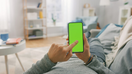 Man at Home Lying on a Couch using Smartphone with Green Mock-up Screen, Doing Swiping, Scrolling Gestures. Guy Using Mobile Phone, Internet Social Networks Browsing. Point of View Shot.