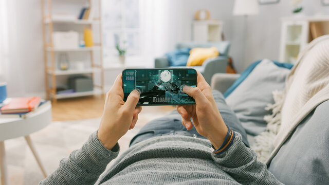 Man at Home Lying on a Couch using Smartphone, Holds it Horizontally in Landscape Mode. He is Playing First Person Shooter Video Game. Point of View Camera Shot.