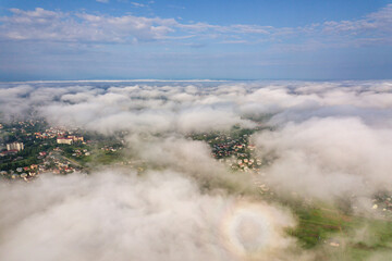 Aerial view of white clouds above a town or village with rows of buildings and curvy streets between green fields in summer. Countryside landscape from above.