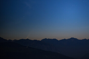 Paesaggio montano alle prime luci dell’alba con nel cielo la cometa C2020 F3 NEOWISE che spunta da dietro le cime dei monti.