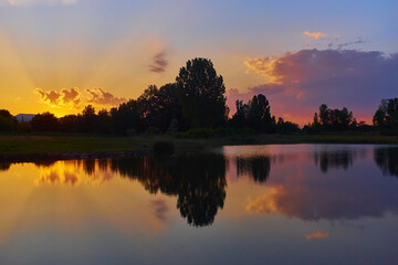 Silhouettes of trees, sunrays and reflections on calm lake water at sunset with vivid yellow, red, blue and purple colors