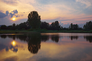 Silhouettes of trees and reflections on calm lake water at sunset with vivid yellow, red, blue and purple colors