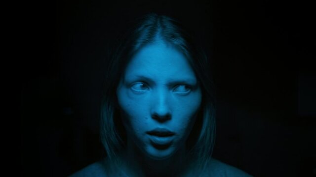 Portrait woman scared face in blue light on black background, grabs head. Woman scared eyes looks camera