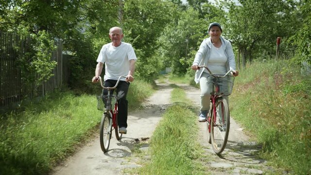 family walk, elderly husband and his beloved old wife enjoy bike ride together on rural road amid green trees on sunny summer day