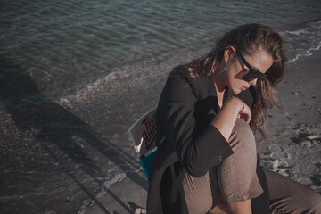 young brunette with long hair in a dark jacket and trousers in sunglasses with a chair on the seashore