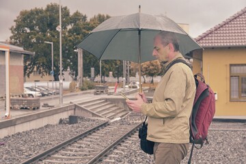 Portrait of a mature traveller texting on smartphone with an umbrella standing on the platform of a train station. Unfinished railway line. When the train arrives.. Funny concept of vain situation