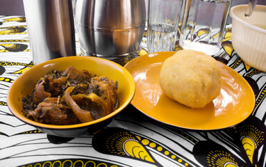 A delicious meal of Nigerian Garri or Eba and Vegetable soup containing assorted meat and fish served in an orange colored bowl and plate on a colorful white and yellow African pattern table cloth