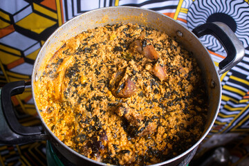 A pot of tasty Egusi soup cooked with assorted meat and dried fish. This is a traditional Nigerian soup ready to be served. A colorful white and yellow pattern cloth is shown in the background