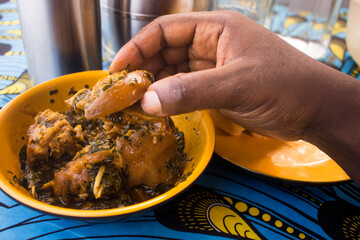 Eating a tasty piece of meat from an orange bowl of Nigerian Vegetable soup containing assorted meat and fish served on a colorful blue and yellow African pattern table cloth