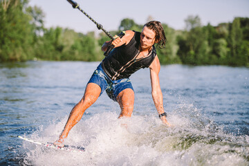 Fototapeta premium Young athletic man with long hair wakesurfing on waves of river in sunny summer weather. Ttheme outdoor activities in summer. Water sports wakesurf on the board. sliding wakeboarder in water splash