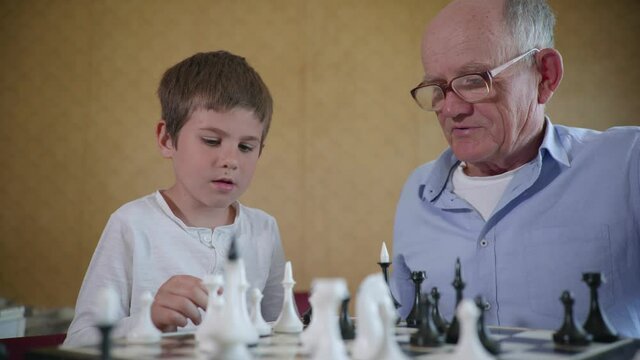 educational games, cute merry male child having fun with his elderly grandfather in glasses for sight in chess sitting at a table in the room during family vacations