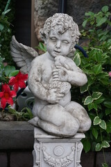 Fototapeta premium Statue of a cupid, angel holding a doll in a flowered garden 1 花の咲いた庭の中にある人形を抱えたキューピッド、天使の像 1