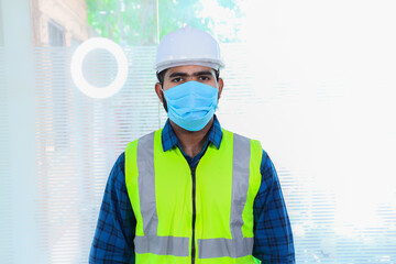 Young engineer wearing mask, closeup of beard man wearing blue shirt with yellow vest and ywhite helmet, back to work after lockdown ends due to covid-19 pandemic, new normal lifestyle