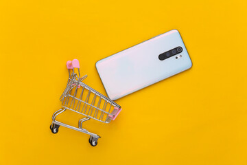 Mini shopping trolley with smartphone on yellow background. Top view