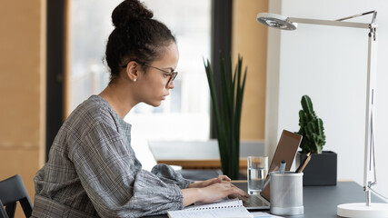 Focused biracial young woman in glasses sit at desk look at laptop screen work consulting client...