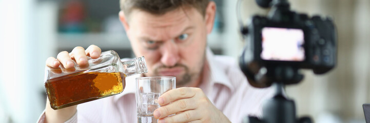 Portrait of man pouring whiskey into glass from bottle. Guy looking drunk and filming on videocamera. Unbutton shirt and bruise under eye. Production on camera