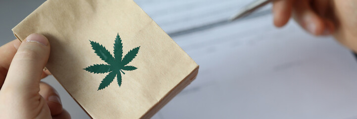 Close-up of person holding package with cannabis. Patients hand signing paper at doctors office. Meds for therapy. Traditional medicine and healthcare concept