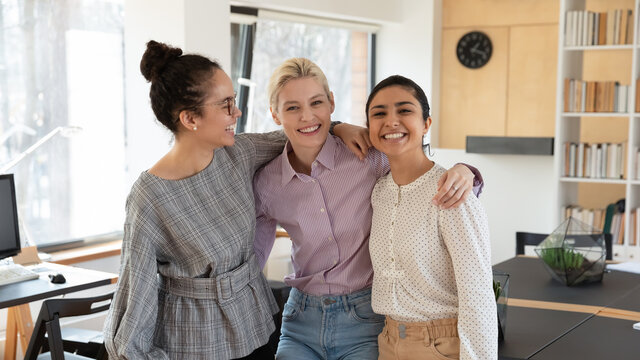 Portrait of smiling young multiracial female colleagues hug posing in modern office together, happy millennial diverse multiethnic women employees embrace at workplace, show friendship and unity