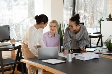 Smiling young multiracial female colleagues laugh working together on laptop in shared office, happy millennial diverse women coworkers cooperate using computer, have fun joke discussing ideas
