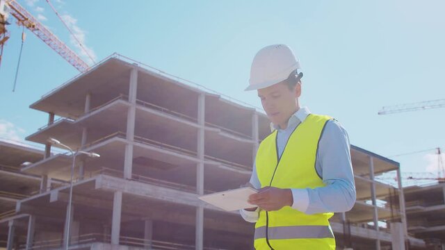 Professional builder standing with tablet computer in front of construction site. Foreman in helmet and vest. Office building and crane background. Business, real estate and investment.