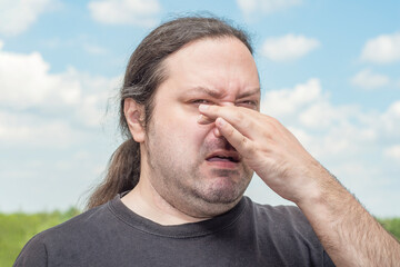 Obraz premium An ordinary middle-aged adult man with long tail hair pinches his nose with two fingers from a bad smell. Man on a light background.