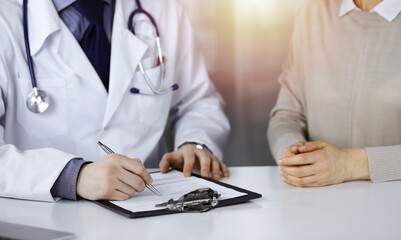 Unknown male doctor and patient woman discussing something while sittingin a darkened clinic and using clipboard, glare of light on the background