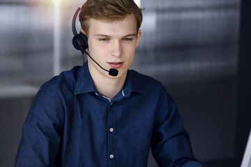 Young blond businessman using headset and computer in a darkened office, glare of light on the background. Startup business means working hard