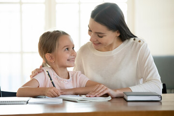 Smiling mother and adorable little daughter studying at home together, pretty preschool girl...