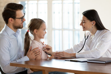 Smiling female pediatrician examining cute little girl sitting on father laps lungs or heart, using stethoscope, doctor gp doing medical checkup in hospital, children healthcare and insurance