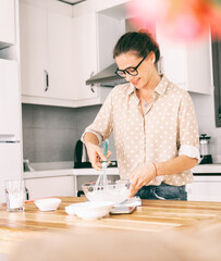 Beautiful young woman in glasses prepares dessert in the kitchen, whips squirrels and dough and smiles happily