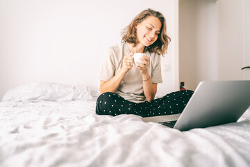 Beautiful young woman enjoying a morning coffee on the bed in front of a laptop, happy smile on his face