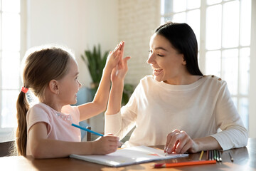 Happy young mother and little daughter giving high five close up, having fun, drawing colorful pencils, sitting at desk together, smiling mum and preschool girl enjoying leisure time at home