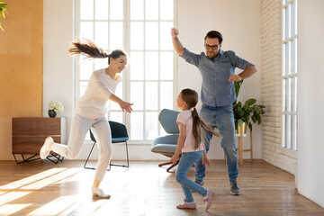 Excited married couple man and woman with little daughter dancing to favorite music in living room,...