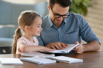 Obraz premium Smiling father helping little daughter with school homework, preparing for lesson, studying at home, sitting at work desk, caring dad checking tasks, reading book, homeschooling concept