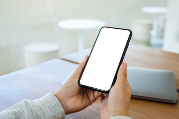 Mockup image of a woman holding mobile phone with blank white desktop screen with laptop on wooden table