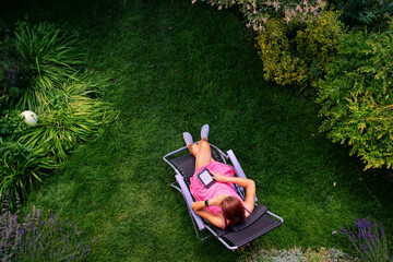 a woman in a red dress lies on a sun lounger on the grass and reads on an ebook reader