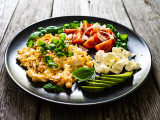 Delicious breakfast - scrambled eggs with fried sausages, cottage cheese, and avocado served on black plate on wooden table
