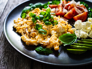 Delicious breakfast - scrambled eggs with fried sausages, cottage cheese, and avocado served on black plate on wooden table
