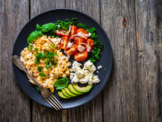 Delicious breakfast - scrambled eggs with fried sausages, cottage cheese, and avocado served on black plate on wooden table
