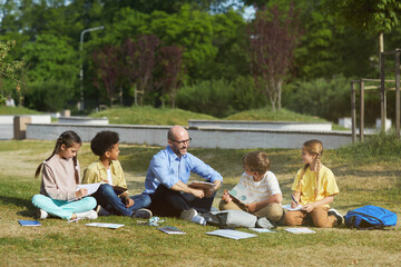 Full length portrait of smiling male teacher talking to group of children while sitting on green grass and enjoying outdoor class, copy space