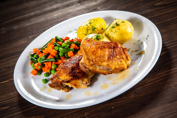 Roasted chicken thighs with boiled carrots, peas and potatoes on wooden background