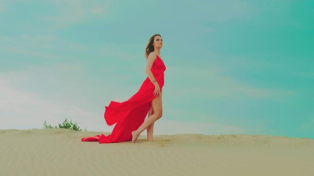 Passionate charming stylish woman posing model on backdrop sandy beach, oasis green coniferous trees, blue hot sky desert. Sexy bare legs. Girl brunette loose hair. Long red silk dress fluttering wind