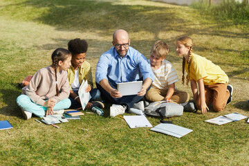 Full length portrait of smiling male teacher talking to group of children while sitting on green...