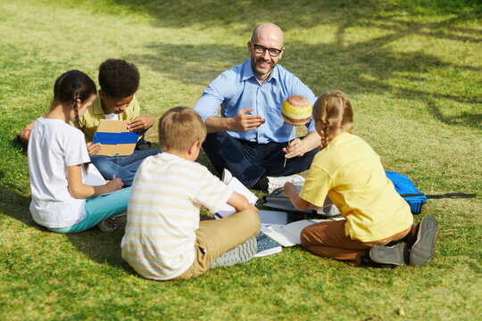 High angle portrait of bald male teacher pointing at planet model and smiling while talking to group of children during outdoor class in sunlight, copy space