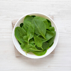 Fresh Baby Spinach in a white bowl on a white wooden background, top view. Overhead, from above, flat lay.