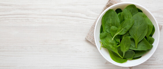 Fresh Baby Spinach in a white bowl on a white wooden background, top view. Overhead, from above, flat lay. Space for text.