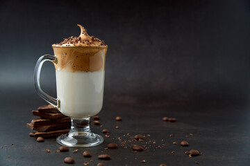 Glass of trendy fluffy creamy dalgona or whipped instant coffee with milk, coffee beans and chocolate on the dark background. New popular food and drink trend concept. Copy space.