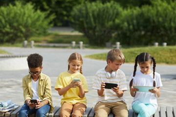 Front view at multi-ethnic group of children using tablets and smartphones while sitting in row outdoors in sunlight, copy space