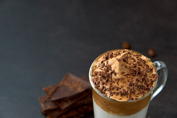 Glass of trendy fluffy creamy dalgona or whipped instant coffee with milk, coffee beans and chocolate on the dark background. New popular food and drink trend concept. Copy space.